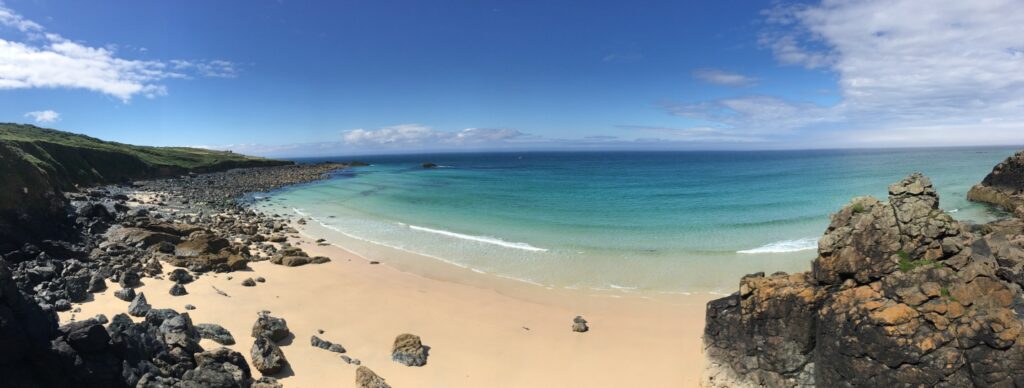 a panoramic view of beach and sea taken from the clifftop. The sea is jade green and some light cirus clouuds break up an otherwise deep blue sky.